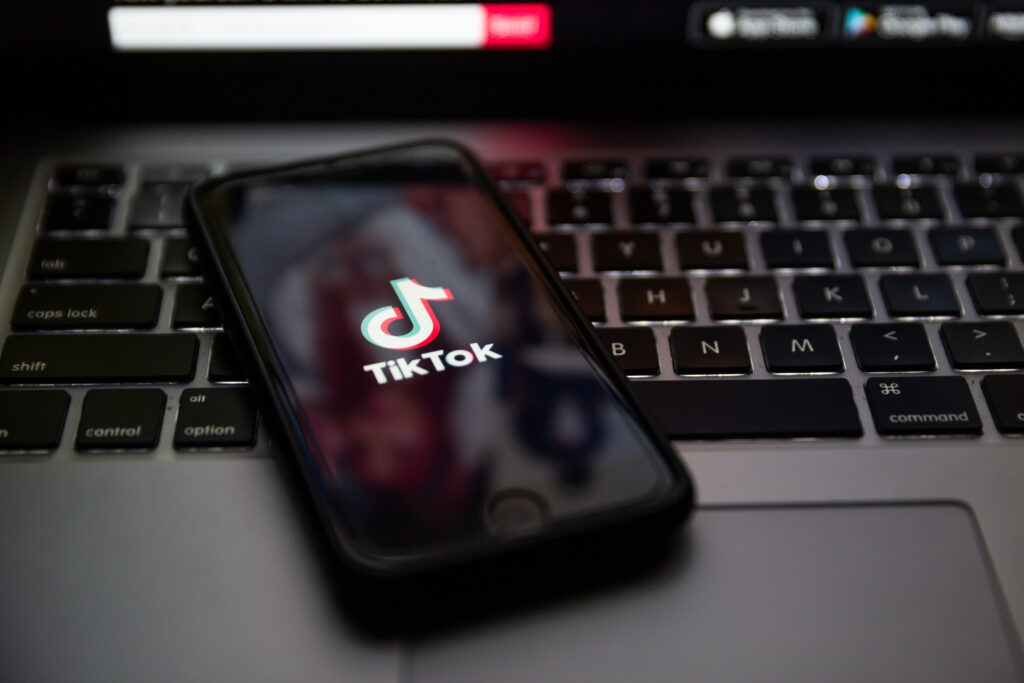 TikTok brand logo on the screen of Apple iPhone 7 in the evening. Keyboard of Macbook Pro in background.